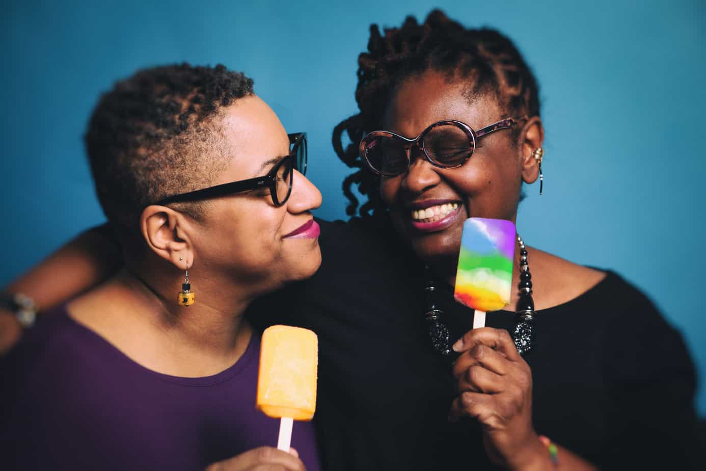 two women hugging, holding brightly colored desserts on a stick.